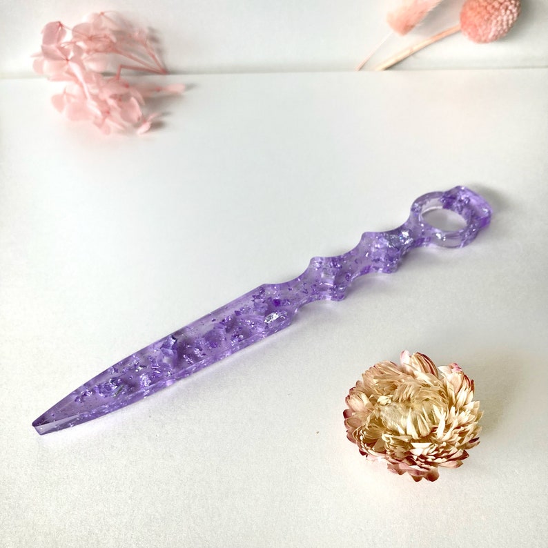 Op de afbeelding: Een paars, doorschijnend, decoratief mes met zilveren glitter ingebed in het plastic. Het mes heeft een unieke, gebogen handgreep met een gat in het midden.