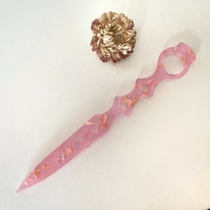Op de afbeelding: Een roze, doorschijnend mes met een decoratief handvat en gouden vlokken. Het mes heeft een puntige punt en een afgerond handvat met een gat in het midden.