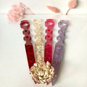 May include: Four decorative hair sticks with geometric designs. The sticks are made of clear resin with gold glitter and come in red, pink, and purple.