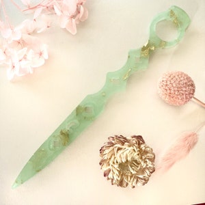 Op de afbeelding: Een lichtgroen, decoratief dolk met gouden vlokken ingebed in de hars. De dolk heeft een afgeronde handgreep met een gat in het midden. De dolk ligt op een wit oppervlak.