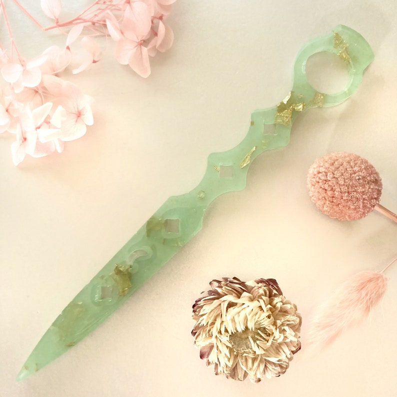 Op de afbeelding: Een lichtgroen decoratief dolk met gouden vlokken. De dolk heeft een afgeronde handgreep met een gat in het midden. Het blad is lang en dun met een puntige punt.