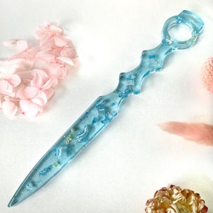 May include: A light blue resin letter opener with a decorative handle and embedded glitter. The handle is shaped like a sword with a rounded tip.