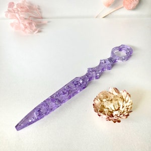 Op de afbeelding: Een paarse brievenopener met een decoratief handvat en zilveren glitter ingebed in de hars. De brievenopener ligt op een wit oppervlak.