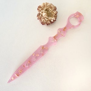 Op de afbeelding: Een roze harsdolk met gouden vlokken en een ringvormig handvat. De dolk heeft een decoratief ontwerp met uitsparingen en is ongeveer 25 cm lang.