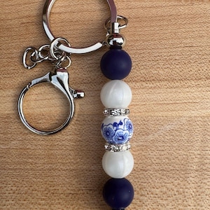 Op de afbeelding: Een zilveren sleutelhanger met een kralen bedel. De bedel heeft marineblauwe, witte en bloemmotief kralen. De kralen zijn geaccentueerd met zilveren strass spacers. De sleutelhanger heeft een karabijnhaak.