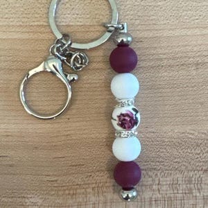 Op de afbeelding: Een zilveren sleutelhanger met een karabijnhaak en een sleutelring. De bedel heeft een reeks kralen: twee bordeauxrode, twee witte en één witte kraal met een bloemenontwerp. De kralen zijn geregen met zilverkleurige metalen accenten.