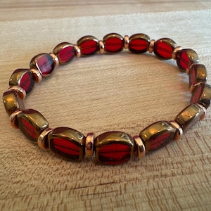 May include: A beaded bracelet featuring red and gold-toned oval beads. The bracelet has a stretch design, with each bead separated by a small, copper-colored spacer. The beads have a glossy finish, reflecting light.