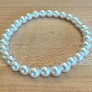 Op de afbeelding: Een witte parelarmband. De armband is gemaakt van ronde, iriserende parels die aan een elastische band zijn geregen. De parels hebben een subtiele glans en zijn in een cirkelvorm gerangschikt. De armband wordt gepresenteerd op een houten oppervlak.