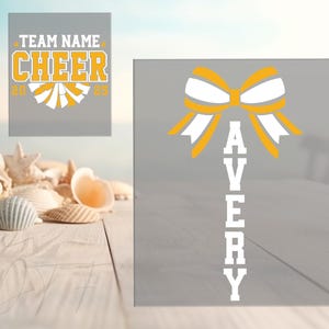 May include: Two grey rectangular signs with cheerleading graphics. One sign has the text "TEAM NAME CHEER 2020" in yellow and white. The other sign has a yellow and white bow graphic with the name "AVERY" in white.