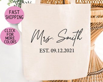 May include: A white canvas tote bag with black text that reads "Mrs. Smith EST. 09.12.2021". The bag is sitting on a wicker chair.