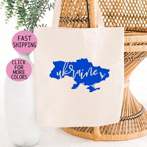 Könnte beinhalten: Eine weiße Stofftasche mit einer blauen Umrandung der Ukraine und dem Wort "ukraine" in weißer Schrift. Die Tasche liegt auf einem Korbstuhl.