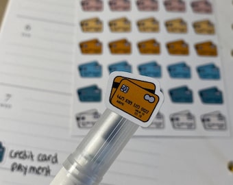 May include: A white pen with a clear plastic barrel is holding a sticker of a credit card. The sticker is yellow and orange with the text "4623 8399 5203 0027" on it. The sticker is on a white planner page with a section labeled "Credit card payment".