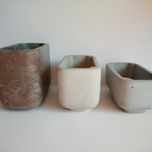 Può includere: Tre vasi rettangolari in cemento di diverse dimensioni. Il vaso più grande è marrone con venature verdi. I due vasi più piccoli sono di colore grigio chiaro.