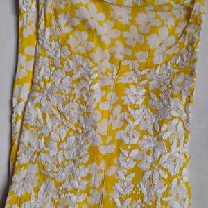 May include: A yellow tunic with a white floral print and embroidered detailing. The tunic features a round neckline and a central slit. The fabric is lightweight, ideal for warmer weather. The embroidery is in a floral and leaf pattern.