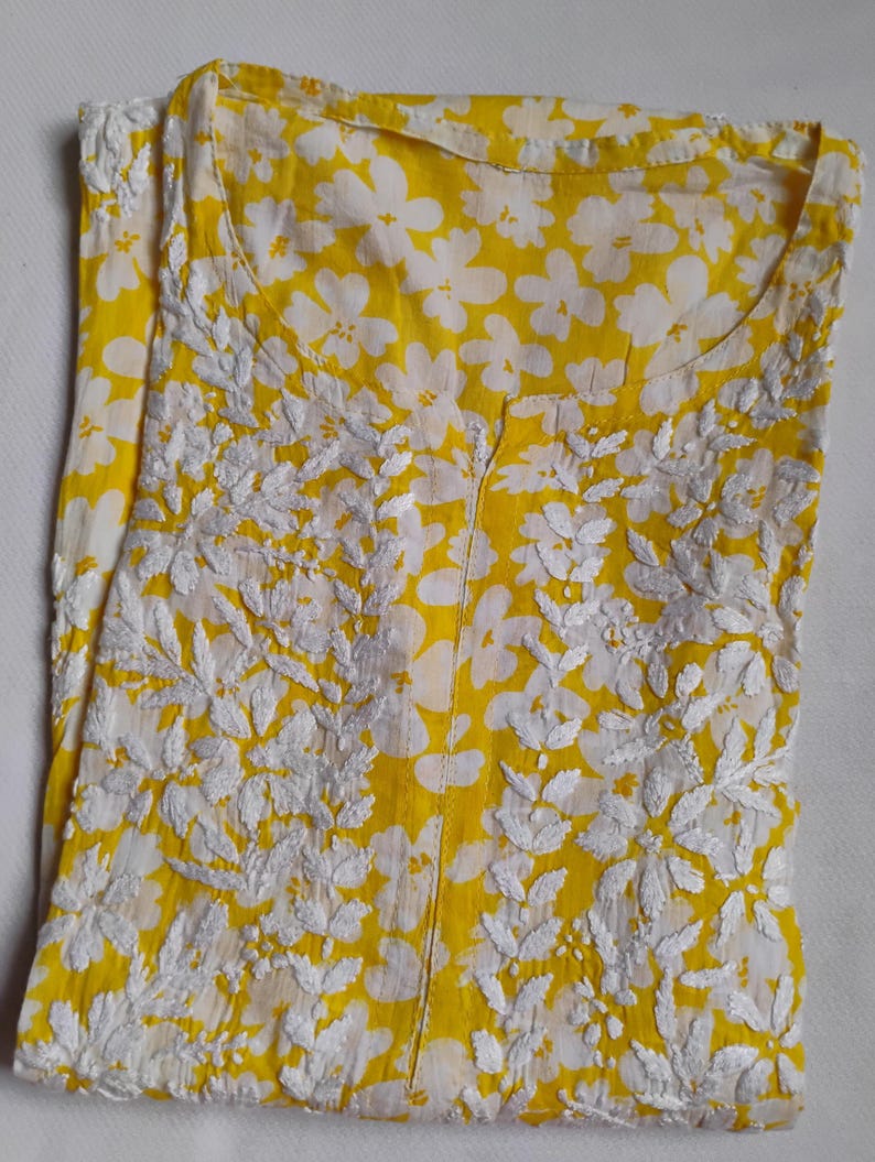 May include: A yellow tunic with a white floral print and embroidered detailing. The tunic features a round neckline and a central slit. The fabric is lightweight, ideal for warmer weather. The embroidery is in a floral and leaf pattern.