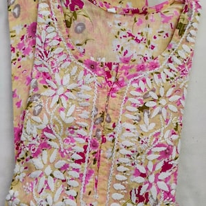 May include: A light yellow floral print tunic with white embroidered floral details and a pink floral pattern. The tunic has a round neckline and a button-up front. The fabric appears to be lightweight and suitable for warm weather.