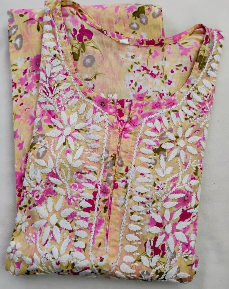 May include: A light yellow floral print tunic with white embroidered floral details and a pink floral pattern. The tunic has a round neckline and a button-up front. The fabric appears to be lightweight and suitable for warm weather.
