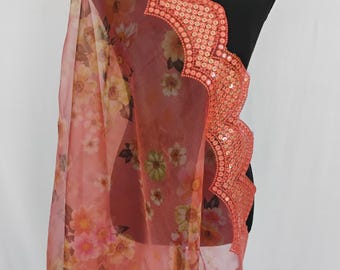 May include: A sheer, coral-colored scarf with a floral pattern. The scarf has a scalloped edge embellished with sequins. The floral design includes pink, yellow, and green flowers. The scarf is draped over a black mannequin.