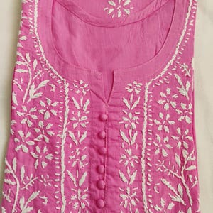 May include: A pink tunic with white floral embroidery. The tunic features a round neckline, a button-down front, and detailed white stitching along the collar, front, and sleeves. The fabric appears to be cotton or a similar lightweight material.