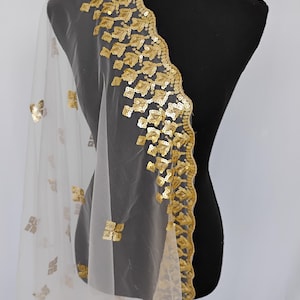 May include: A sheer white fabric with gold sequin embellishments. The fabric is draped over a black mannequin, showcasing a detailed border of gold sequins and scattered gold sequin accents.