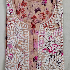 May include: A folded peach-coloured tunic with floral embroidery and a floral print. The tunic features white embroidered floral designs around the neckline and down the front. The floral print includes shades of pink, red, and purple.