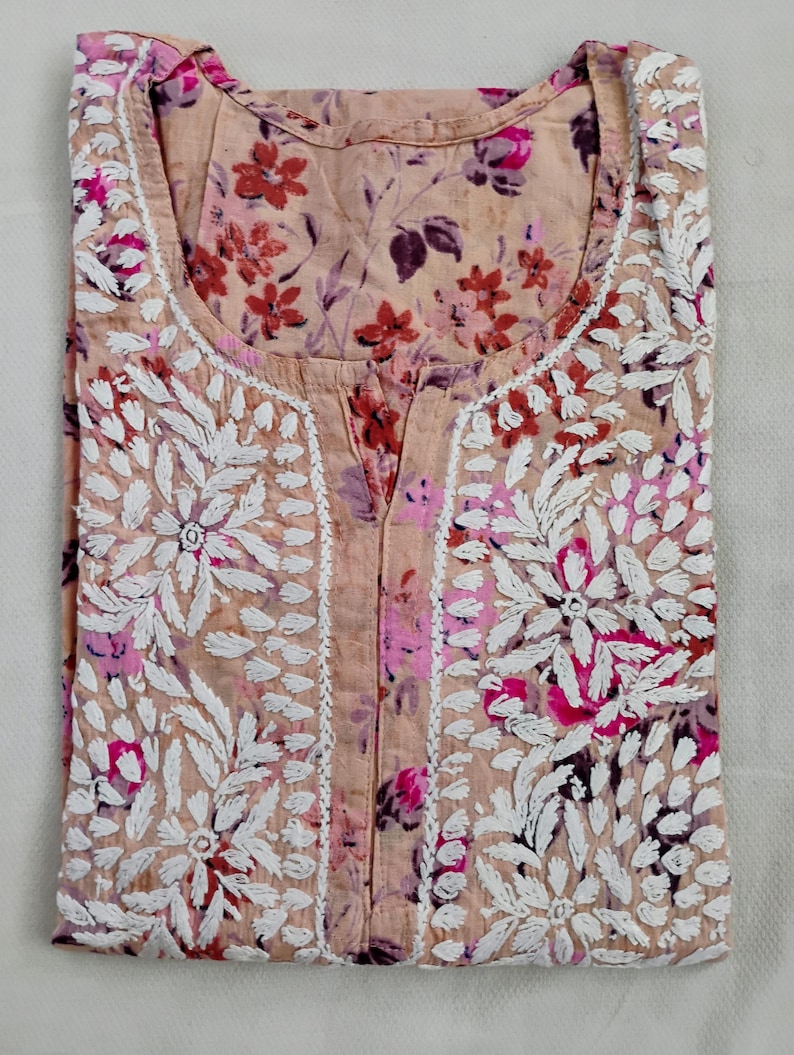May include: A folded peach-coloured tunic with floral embroidery and a floral print. The tunic features white embroidered floral designs around the neckline and down the front. The floral print includes shades of pink, red, and purple.