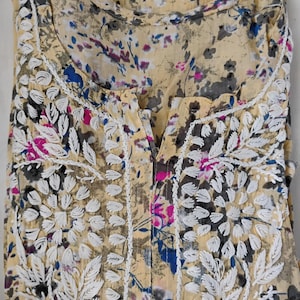 May include: A folded, pale yellow blouse with a floral pattern in blue, pink, and grey. The blouse has white embroidered floral details on the neckline and front. The fabric looks lightweight and slightly sheer.