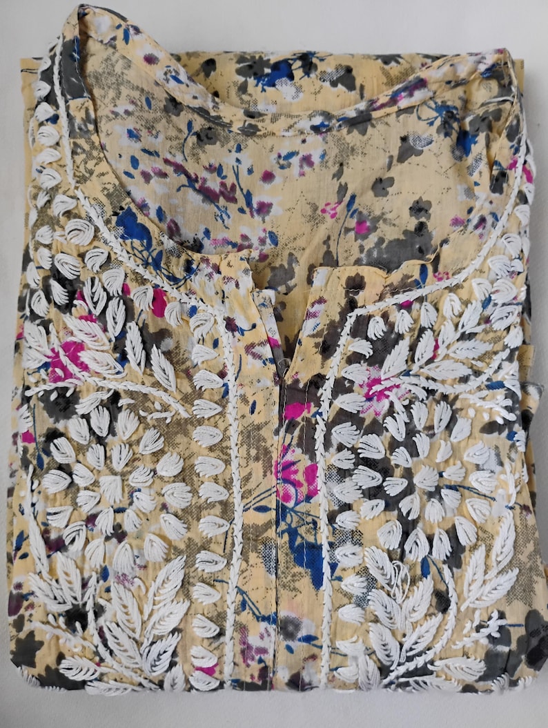 May include: A folded, pale yellow blouse with a floral pattern in blue, pink, and grey. The blouse has white embroidered floral details on the neckline and front. The fabric looks lightweight and slightly sheer.