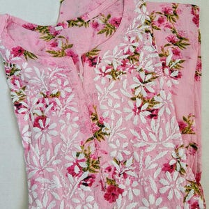 May include: A light pink, two-piece outfit featuring a floral print and white embroidered details. The top has a round neckline and a split front. The fabric is a soft, lightweight material, perfect for warm weather. The floral pattern includes pink and red flowers with green leaves.