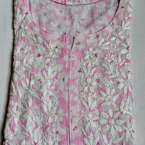 May include: A folded, pink and white floral embroidered tunic. The tunic has a round neckline and a split front. The fabric is a light pink with a white floral pattern. The embroidery is white with small pink accents.