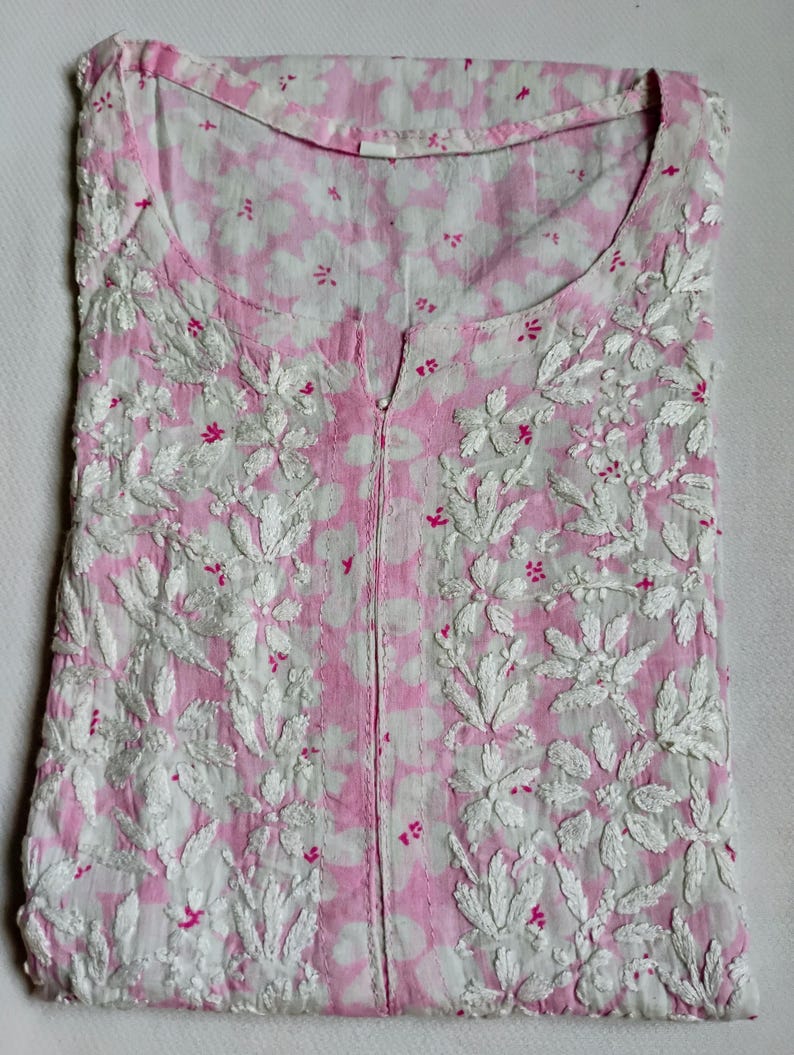 May include: A folded, pink and white floral embroidered tunic. The tunic has a round neckline and a split front. The fabric is a light pink with a white floral pattern. The embroidery is white with small pink accents.