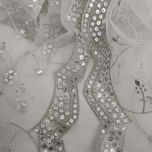 May include: Close-up of a white sheer fabric with silver sequin and floral embroidery. The fabric has a wavy, embellished border with rows of silver sequins. Delicate silver thread outlines floral patterns.