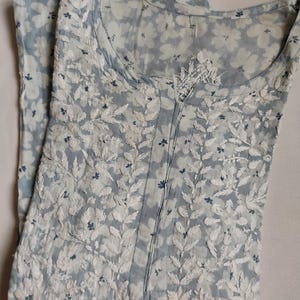 May include: A pale blue dress featuring a floral print and white embroidered leaf patterns. The dress has a round neckline and a buttoned front. The fabric is lightweight, ideal for warmer weather.