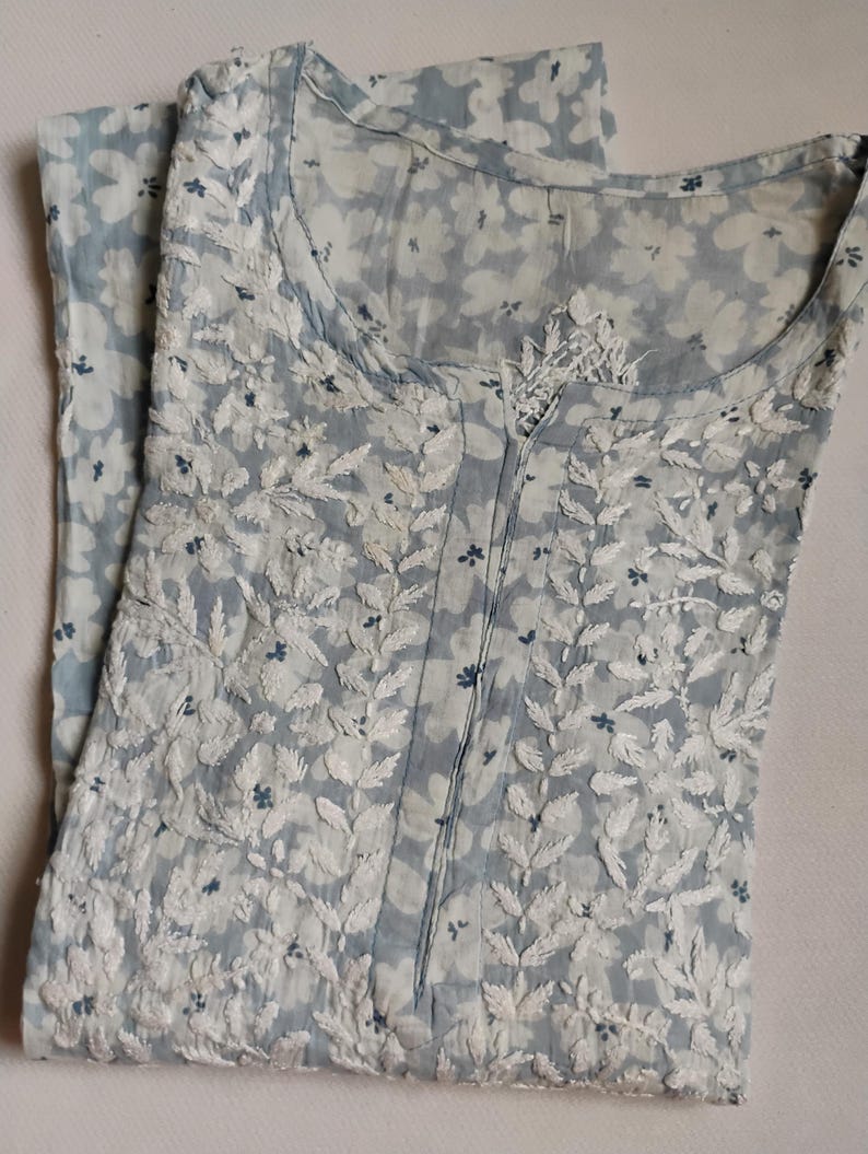 May include: A pale blue dress featuring a floral print and white embroidered leaf patterns. The dress has a round neckline and a buttoned front. The fabric is lightweight, ideal for warmer weather.