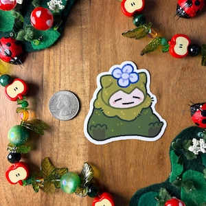 Pu&ograve; includere: Un adesivo raffigurante un personaggio dei cartoni animati in tonalit&agrave; verdi con un fiore blu sulla testa. L'adesivo &egrave; su una superficie di legno, circondato da ornamenti decorativi a forma di mela e coccinella. &Egrave; presente anche una moneta americana.