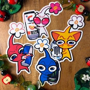 May include: A collection of colorful stickers featuring cartoon-like creatures holding energy drink cans. The stickers include red, white, yellow, and blue characters with large eyes and flower accents. The drinks include Red Bull, Monster, and other brands.