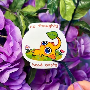 May include: A sticker depicting a yellow cartoon creature in a fishbowl, with the phrases "no thoughts" and "head empty". The creature has blue eyes and a green leaf. The sticker is set against a backdrop of purple flowers and green foliage.