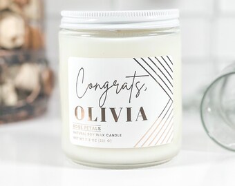 May include: A glass jar candle with a white lid and a label that reads "Congrats, Olivia." The label also includes the words "Rose Petals" and "Natural Soy Wax Candle." The candle weighs 7.6 oz (221 g).