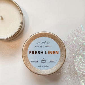 Puede incluir: Una vela de soja mini dorada con una etiqueta blanca que dice "Low Candle Co. MINI SOY CANDLE FRESH LINEN 2 oz/57g 8 hours made with love".