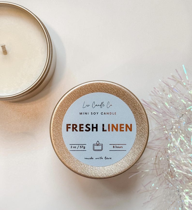 Puede incluir: Una vela de soja mini dorada con una etiqueta blanca que dice "Low Candle Co. MINI SOY CANDLE FRESH LINEN 2 oz/57g 8 hours made with love".