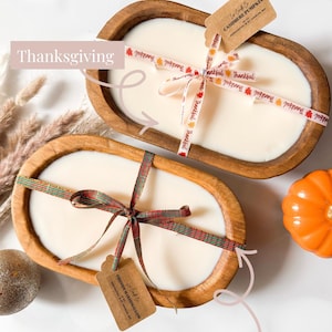 Puede incluir: Dos velas en cuencos de madera ovalados rellenos de cera blanca. La vela superior tiene una cinta con la palabra Thankful y una etiqueta Cashmere Pumpkin. La vela inferior tiene una cinta de cuadros y una etiqueta Fireside Marshmallow.