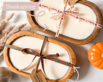 May include: Two oval wooden dough bowl candles filled with white wax. The top candle features a ribbon printed with the word Thankful and a tag labeled Cashmere Pumpkin. The bottom candle has a plaid ribbon and a tag labeled Fireside Marshmallow.