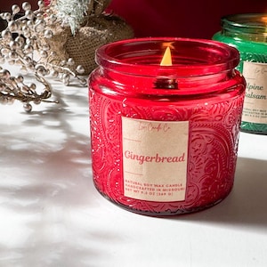 Puede incluir: Una vela encendida en un tarro de cristal rojo con la palabra "Gingerbread" en la etiqueta. La vela pesa 269 g y está hecha a mano en Missouri. Una segunda vela verde es visible en el fondo.