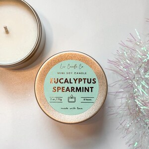 Puede incluir: Una vela en una lata dorada con una etiqueta azul claro que dice "Lu Candle Co. MINI SOY CANDLE EUCALYPTUS SPEARMINT 2 oz/57g 8 hours made with love"