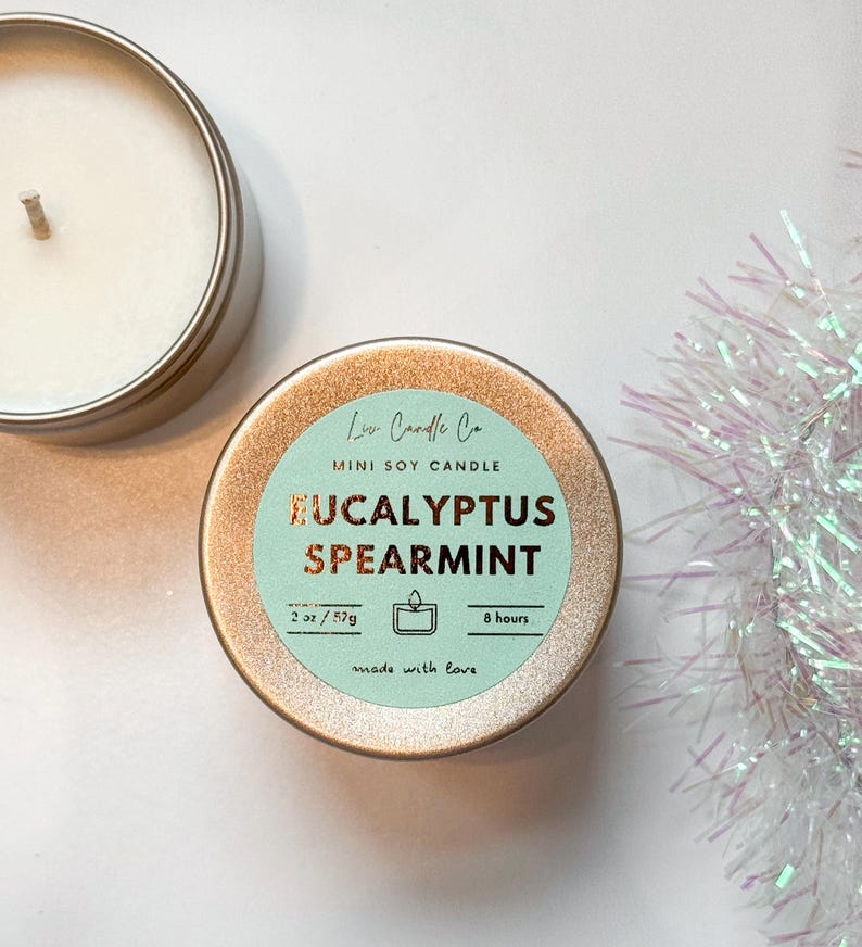 Puede incluir: Una vela en una lata dorada con una etiqueta azul claro que dice "Lu Candle Co. MINI SOY CANDLE EUCALYPTUS SPEARMINT 2 oz/57g 8 hours made with love"