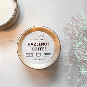 Puede incluir: Una lata de vela de soja dorada en miniatura con una etiqueta blanca que dice "Liv Candle Co. MINI SOY CANDLE HAZELNUT COFFEE 2 oz/57g 8 hours made with love".