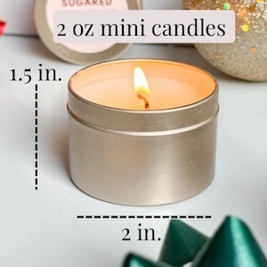 Puede incluir: Una vela peque&ntilde;a en una lata dorada con un interior de cera blanca y una mecha encendida. La vela tiene 5 cm de di&aacute;metro y 3,8 cm de alto. El texto "2 oz mini candles" es visible sobre la vela.