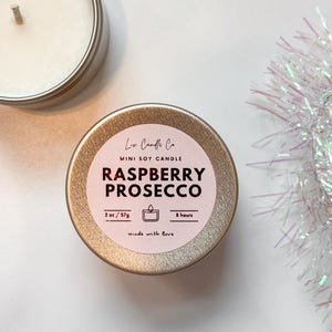 Puede incluir: Una vela en una lata dorada con una etiqueta rosa que dice "Loa Candle Co. Mini Soy Candle Raspberry Prosecco 2 oz/57g 8 hours made with love".