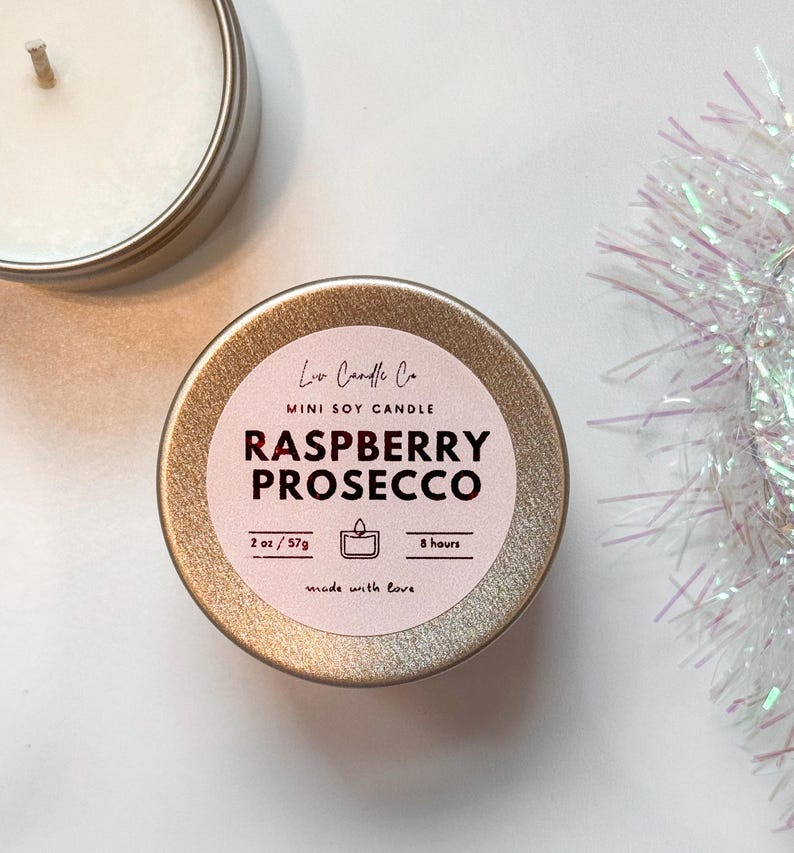 Puede incluir: Una vela en una lata dorada con una etiqueta rosa que dice "Loa Candle Co. Mini Soy Candle Raspberry Prosecco 2 oz/57g 8 hours made with love".