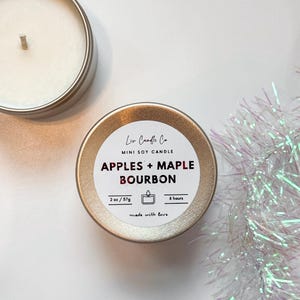 Puede incluir: Una lata de vela de soja dorada en miniatura con una etiqueta blanca que dice "Liv Candle Co. MINI SOY CANDLE APPLES + MAPLE BOURBON 2 oz/57g 8 hours made with love".